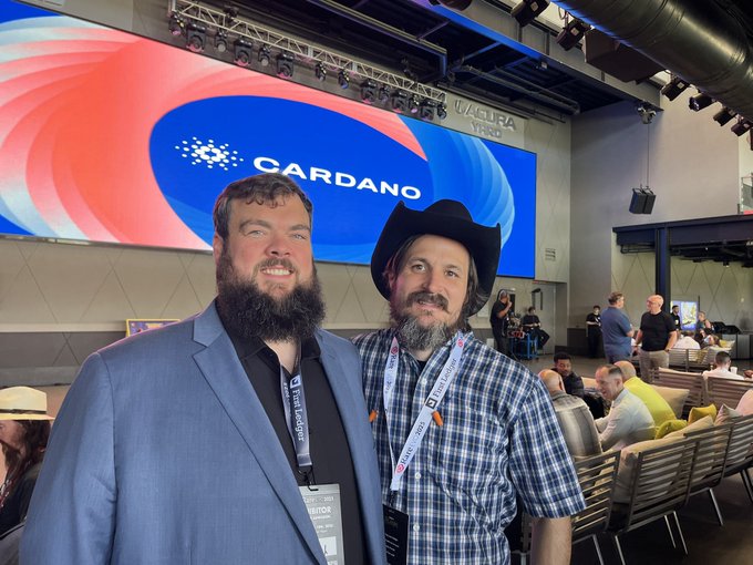 Two bearded men posing for a photo at a Cardano-branded event venue, with one wearing a black cowboy hat and the other in a blue suit jacket.
