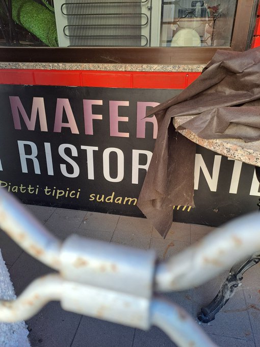 A close-up of a black storefront sign reading 'MAFER RISTOR... Piatti tipici sudam...' partially covered by a brown tarp, with a rusty metal chain lock in the foreground.
