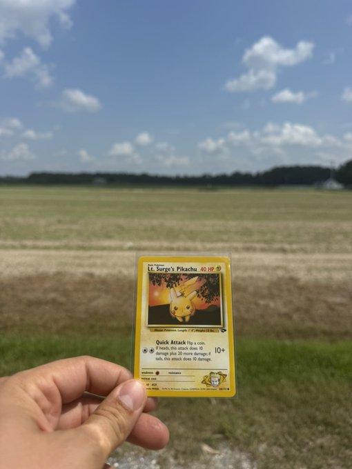 A hand holding up a 'Lt. Surge's Pikachu' Pokemon trading card (60 HP, 1st Edition) against a backdrop of a blue sky with clouds over an open field.