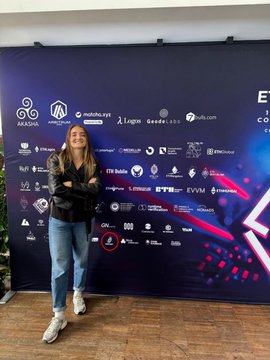 The image features a young woman standing next to a large banner or wall that displays various logos and text, including "Captain Capital" and "@ETHCluj 2026". The woman appears to be posing for a photo with her arms crossed, possibly at an event related to Web3 technology. The background of the image is dark, which contrasts with the bright colors on the banner and makes it stand out more prominently.