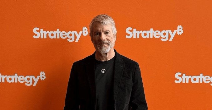 Michael Saylor, the founder of MicroStrategy (now branded as 'Strategy'), stands smiling in front of an orange backdrop featuring the repeated 'Strategy' logo with the Bitcoin 'B' symbol.