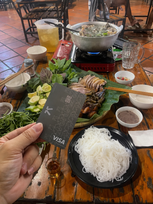A hand holds a black Visa credit card in front of a Vietnamese meal featuring a hot pot, rice noodles, fresh herbs, grilled meat, and dipping sauces on a wooden table.