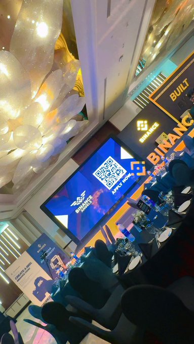 An indoor event space decorated for Binance's 9th anniversary celebration features a stage setup with screens displaying the Binance logo, a QR code, and a "9" graphic, alongside an "APPLY NOW!" banner, complementing the post's tribute to nine years of Binance's innovation and global crypto community building.