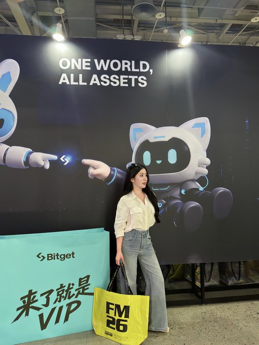 A woman poses in front of a 'Bitget' advertisement banner reading 'ONE WORLD, ALL ASSETS' featuring a cartoon robot mascot, holding a yellow 'FM 26' shopping bag.