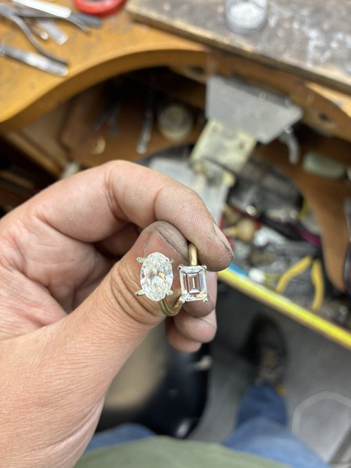 A jeweler's dirty, grease-stained hand holds two diamond engagement rings—an oval cut and an emerald cut—displaying them above a cluttered workbench. The image captures the raw, in-progress craftsmanship of the rings, fitting the poster's anticipation of the finished "two-stone" design with a playful tone.