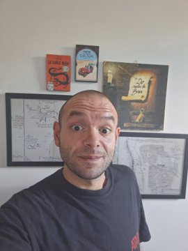 The image features a man with a shaved head, wearing a black t-shirt, standing against a white wall. He has his mouth open wide and appears to be making a funny or surprised face for the camera. The background of the photo includes several framed pictures on the wall behind him, adding an artistic touch to the scene. There is also text visible in the image that reads "Gooooood morning beautieeees! Today you’ll see me posting something I’ve already posted and reposted, but...I added the IMX tag, and the result was...take note: -engagement completely wiped out -the attached video disappears on mobile view (as you can see in the photo)".