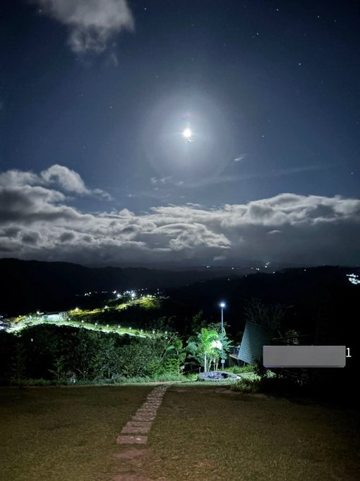 The image features a beautiful night scene with a full moon illuminating the sky above a mountain range, creating an enchanting atmosphere. The moonlight casts a soft glow over the landscape below, highlighting the silhouettes of trees against the dark sky. In the foreground, there is a path leading towards a building or structure that can be seen in the distance. The image also contains text elements such as "Good night" and "@useTria isn't slowing down."
