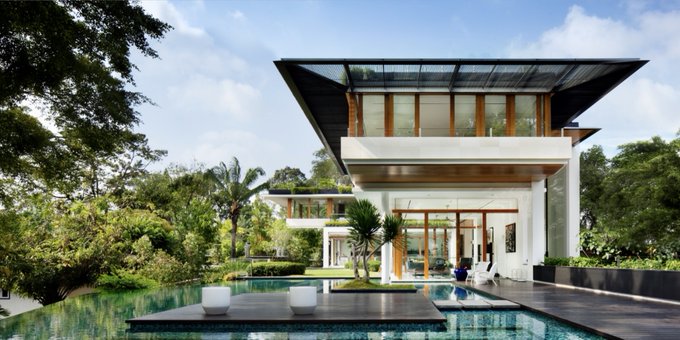 A modern luxury home with large glass windows and a flat, overhanging roof is situated behind a long, dark swimming pool, surrounded by lush tropical greenery.