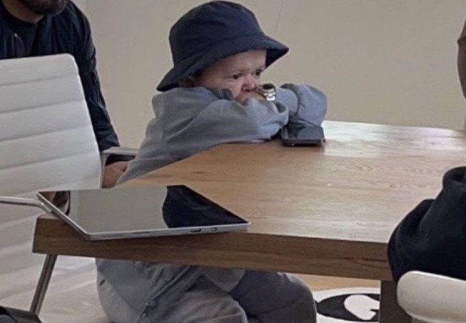 A smug baby wearing a black bucket hat rests their chin on their hands at a conference table, appearing to eye a laptop like a shrewd investor. The image cleverly captures the bitter, knowing smile of someone watching their sold memecoin skyrocket 1000% while they remain on the sidelines.