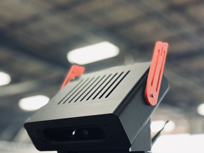 Close-up of a black industrial camera or sensor device with red mounting brackets, viewed from below against a blurred warehouse ceiling with overhead lights.