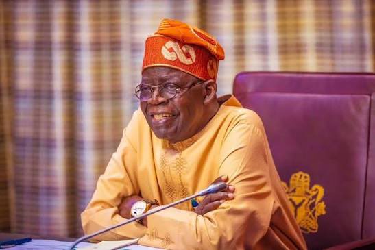 A photograph of Bola Ahmed Tinubu, the President of Nigeria, wearing traditional yellow attire and a red and white cap with the number '67' on it, smiling while seated at a desk.