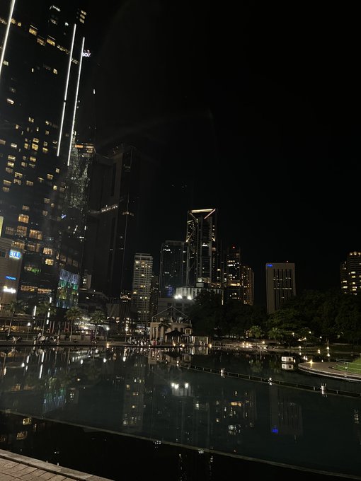 Nighttime photograph of a city skyline with illuminated skyscrapers reflected in a dark pool of water in the foreground.