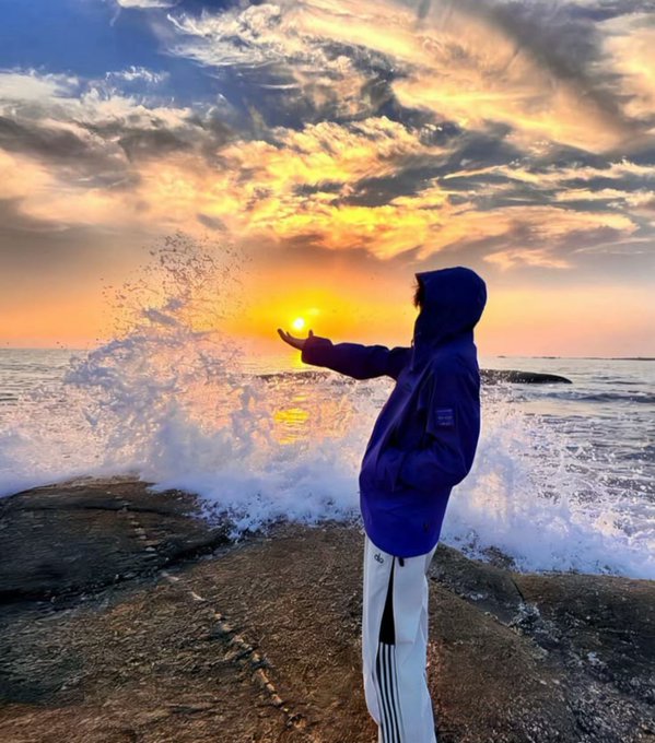 A person in a blue hooded jacket stands on rocky shore, reaching out toward a crashing wave under a dramatic sunset sky.