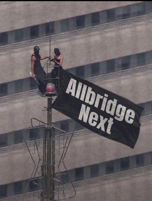 Two people stand on a tower platform next to a large black flag reading 'Millbridge Next' in white text.