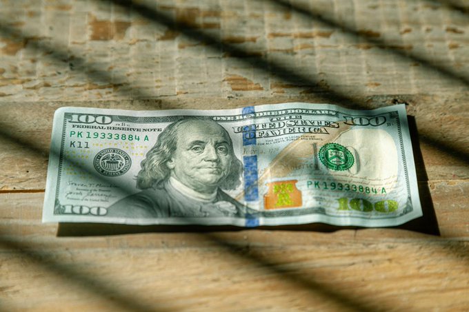 A slightly crumpled $100 U.S. bill resting on a wooden surface, with the serial number 'PK19333884 A' and 'K11' clearly visible.