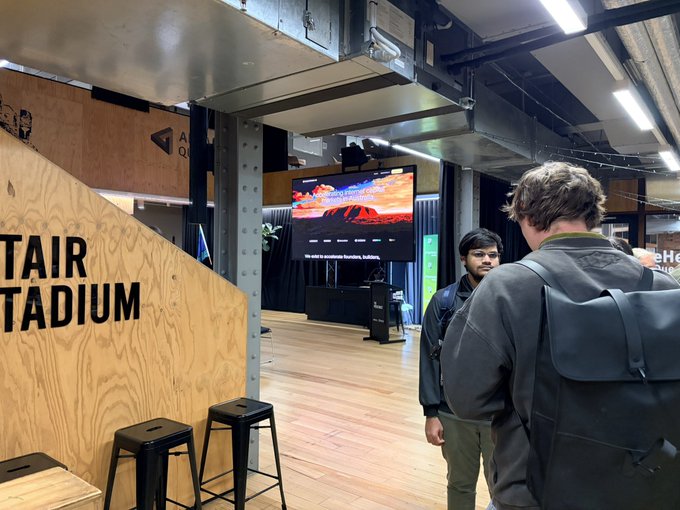 Interior of an event space featuring plywood walls labeled 'STAIR' and 'STADIUM', with attendees gathered around a central presentation screen.