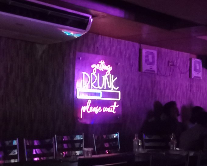The image features a neon sign that says "Getting Drunk Please Wait" written in pink neon letters. The sign is located inside a bar or restaurant with people sitting at tables enjoying their time. There are chairs and dining tables visible, indicating that this place serves food as well. The neon sign seems to be promoting the idea of Proof of Useful Work (PoWH), which is an alternative method for mining cryptocurrencies like Bitcoin. PoWH involves solving math puzzles in exchange for cryptocurrency, making it a more energy-efficient way to mine compared to traditional methods.