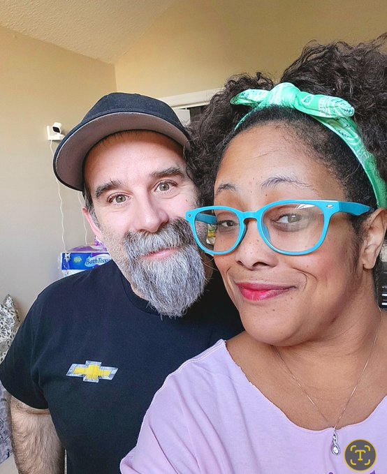 Selfie of a smiling couple - a bearded man wearing a black Chevrolet cap and a woman with curly hair, green headband, and turquoise glasses.