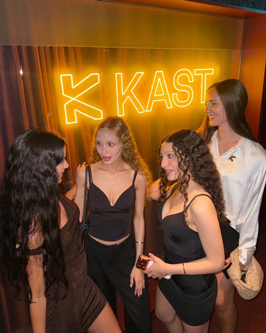 A group of four stylish young women in dark dresses gather and chat beneath a glowing orange "X KAST" neon sign, capturing a fun girls' night out. The vibrant nightlife backdrop perfectly complements the caption's celebration of a memorable evening with friends.