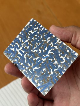 A hand holds a small rectangular object or card decorated with a busy blue, white, and tan camouflage-like pattern over a wooden table.