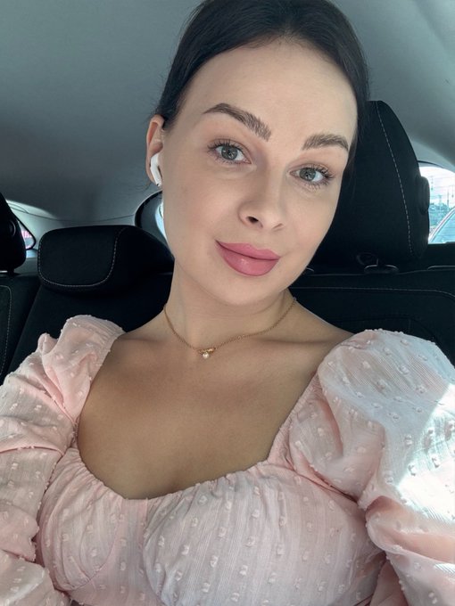 A selfie of a young woman with dark hair wearing a pink textured top and white earbuds, sitting in a car.