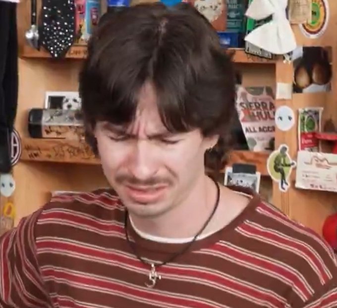 A young man with dark hair and a goatee grimaces with a pained expression, wearing a red and brown striped shirt in front of a wall covered with various stickers and posters.