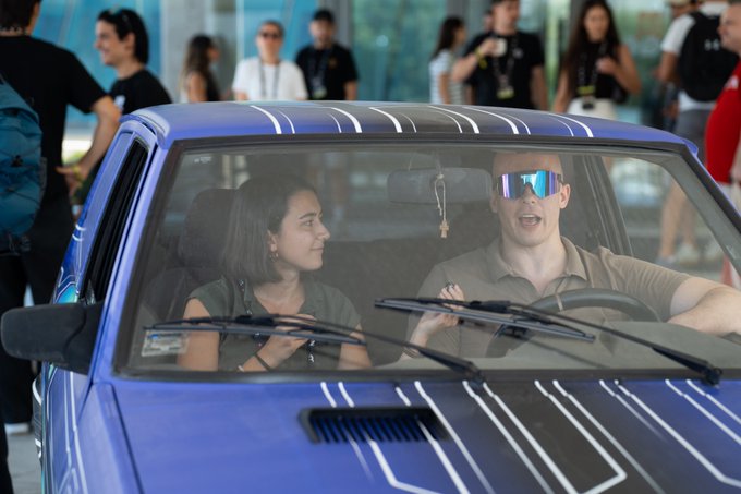 Two people, a woman with dark hair and a man wearing large blue reflective sunglasses, sit inside a blue car with a black patterned hood, appearing to talk while bystanders watch in the background.