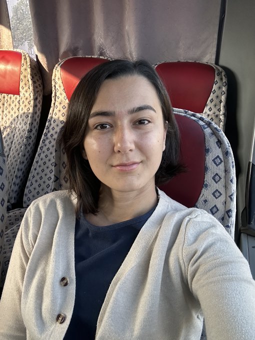 A close-up selfie of a young woman with short dark hair wearing a beige cardigan over a dark blue shirt, sitting in a seat with a patterned red and beige headrest.