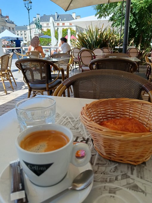 A first-person view at an outdoor café featuring a cup of espresso labeled 'Cellini,' a glass of water, and a wicker basket containing a croissant, with wicker chairs and a city square visible in the background.