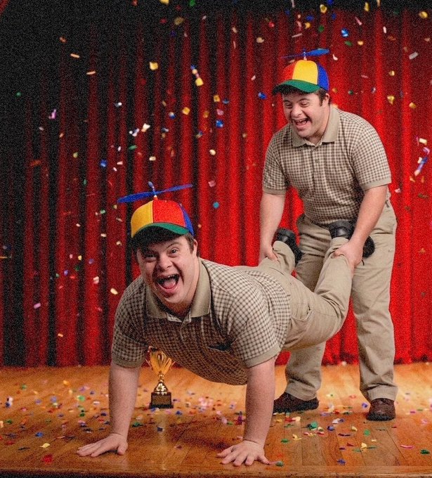 A comedic photograph of two men wearing propeller beanies performing a silly acrobatic stunt on a stage, with confetti falling and a small trophy on the floor.