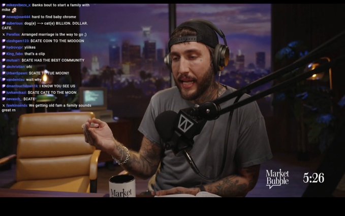 A bearded man wearing headphones and a cap hosts the "Market Bubble" show at timestamp 5:26, speaking into a microphone while gesturing with a pen, as live chat viewers discuss "$CATE" (CATE COIN TO THE MOON) and reference Banks and his new family.