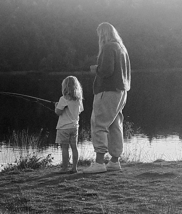 A nostalgic black and white photograph captures a long-haired person and a small child standing by a misty lake, with the child holding a fishing rod.