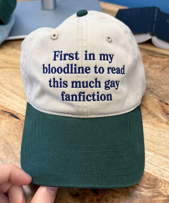 A two-toned beige and green baseball cap with embroidered text reading 'First in my bloodline to read this much gay fanfiction.'