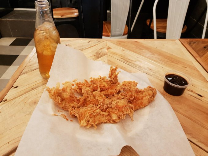 A basket of crispy fried chicken tenders served on parchment paper alongside a small cup of dipping sauce and a glass bottle of iced tea, presented on a wooden restaurant table.