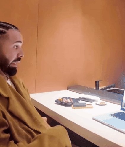 The image shows a man sitting at a desk with an open laptop computer placed on top of him. The man appears surprised or shocked by something happening on his screen. There are also two cell phones visible near the edge of the desk, possibly belonging to the man or someone else in the room.