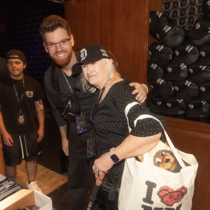 A man in glasses poses with an older woman wearing a black polka-dot top and a "DD" cap; she carries a tote bag with a pixel heart and "I ❤ DD" design.
