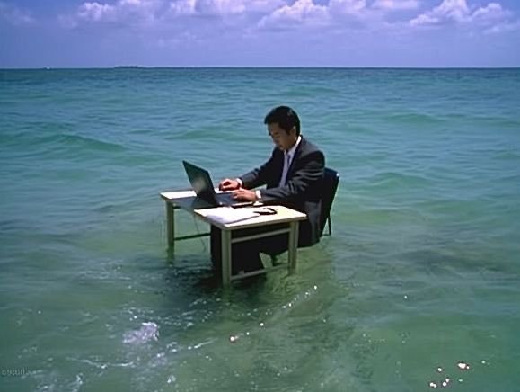 A surreal image of a businessman in a suit working at a desk with a laptop while the furniture is partially submerged in the ocean water.