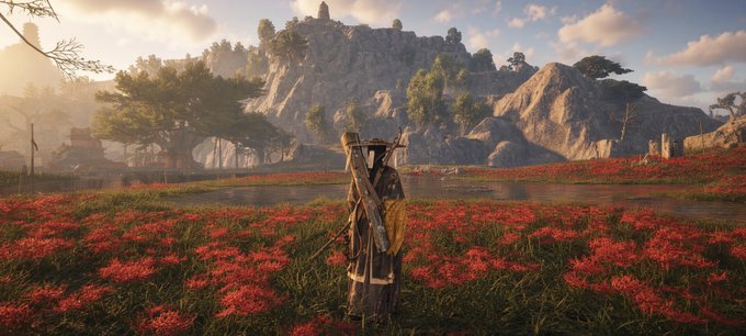 A video game character stands in a field of red flowers facing a lake and mountains at sunset, appearing to be from Ghost of Tsushima.