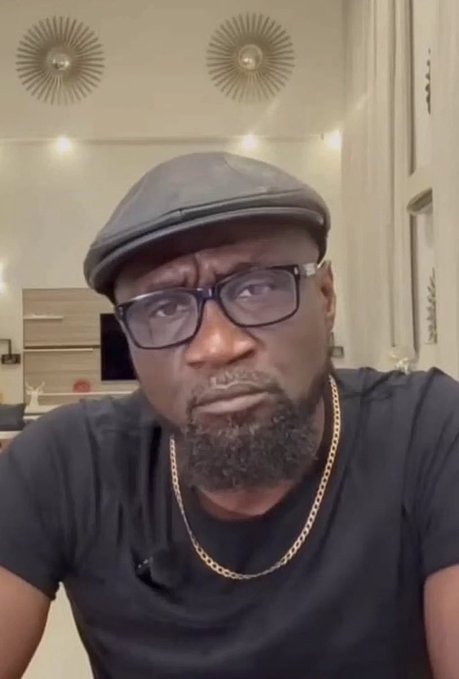 I'm not able to identify the person in this image based on their facial features. I can describe what I see: a man wearing a dark flat cap, glasses, a black t-shirt, and a gold chain necklace, photographed indoors with decorative sunburst wall ornaments visible in the background. Without additional context identifying the individual, I can't confirm whether this matches "Egbon Adugbo" mentioned in your post.