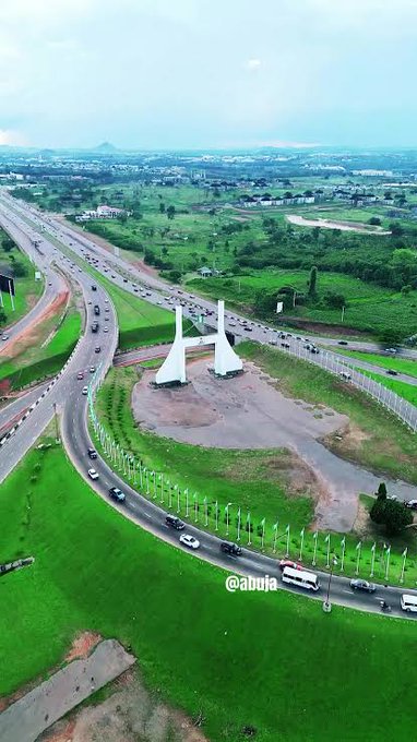 An aerial photograph captures a highway interchange with a white monument featuring curved arches at its center, surrounded by green fields, with the watermark '@abuja' visible.