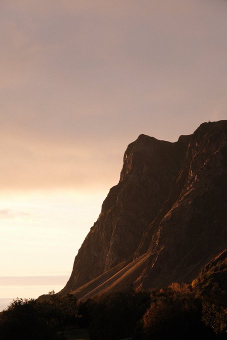 A dramatic sunset or sunrise scene featuring a silhouetted rocky mountain cliff against a soft pastel sky with a view of the ocean below.