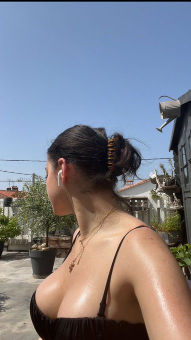 A woman with dark hair pulled back in a claw clip, wearing a black top and wireless earbuds, stands outdoors on a sunny day with potted plants and a residential building visible in the background.