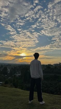 A person silhouetted standing on a grassy hill, gazing at a vibrant sunset sky filled with scattered clouds.