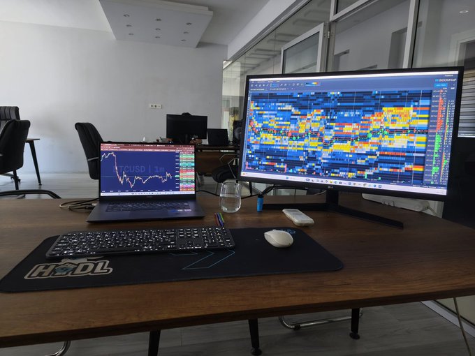 A professional trading workstation with a large curved monitor showing order book heatmap data, a laptop displaying BTCUSD price chart, a keyboard, mouse, and an HODL-branded mouse pad.