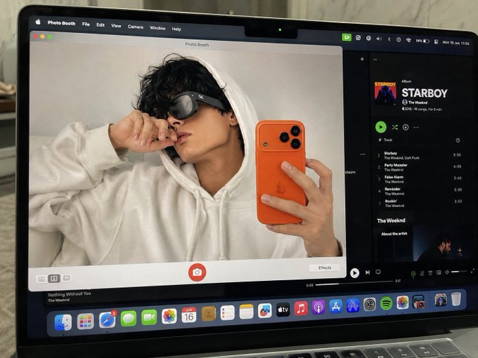 A MacBook screen displaying the Photo Booth app with a selfie of a person wearing sunglasses and a white hoodie holding an orange iPhone 17 Pro, with Spotify playing 'STARBOY' by The Weeknd in the background.