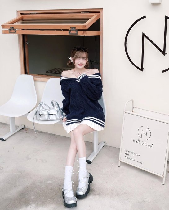 A young woman with light brown hair wearing sunglasses, a navy blue off-shoulder sweater, white skirt, and platform shoes, sitting on a white chair outside 'nubi island' cafe.
