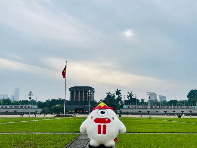 A white one-eyed plush character wearing a red headband stands on the lawn of Ba Đình Square in Hanoi, with Vietnam's flag-raising ceremony, Ho Chi Minh Mausoleum, and banners reading "NGHĨA VIỆT NAM MUÔN NĂM!" and "CHỦ TỊCH HỒ CHÍ MINH VĨ ĐẠI SỐNG MÃI TRONG SỰ NGHIỆP CỦA CHÚNG TA!" visible in the background, capturing a festive moment ahead of Vietnam's National Day on September 2nd.