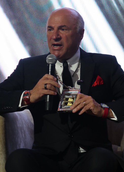 Kevin O'Leary, dressed in a black suit with a red pocket square, speaks into a microphone while proudly displaying his rare one-of-one Shohei Ohtani trading card in a protective case. The image captures the moment he publicly unveils the prized $11 million card, with his smartwatch and red bracelet visible on his wrists.