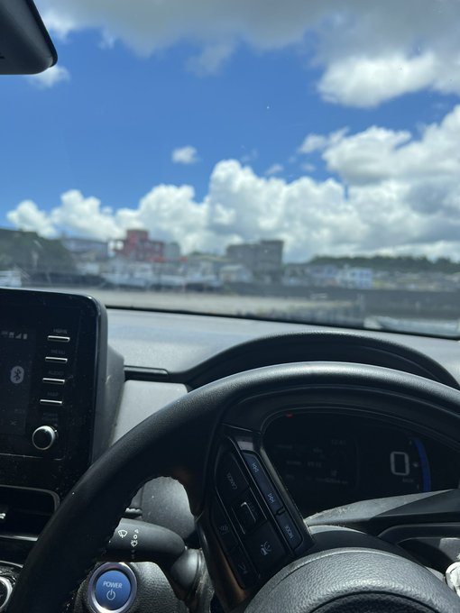 A view from inside a car shows the steering wheel, dashboard, and a scenic view of a coastal town under a bright blue sky with scattered clouds.