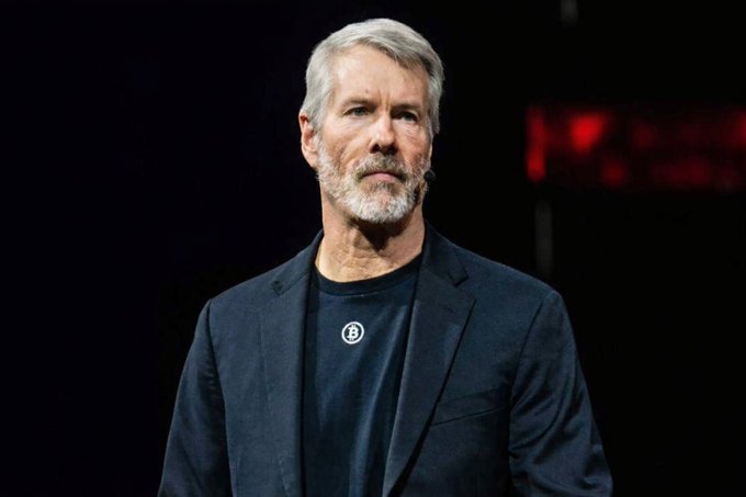A man with gray hair and a beard, wearing a dark blazer over a black t-shirt featuring a Bitcoin logo, stands against a dark background—likely Michael Saylor, executive chairman of Strategy (MSTR). The Bitcoin-themed image reinforces the post's bullish message about holding $MSTR stock despite the temporary dip.